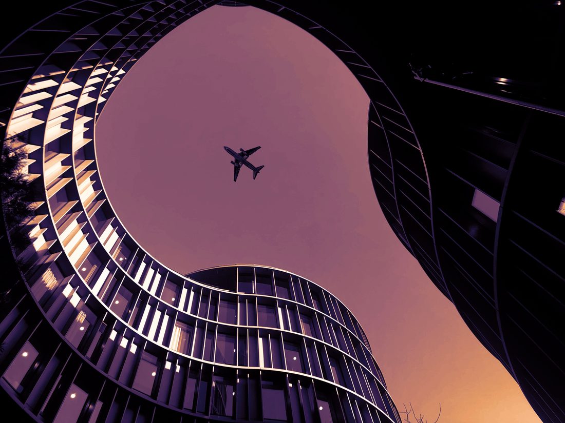 An aircraft passing overhead between curved glass buildings at dusk.
